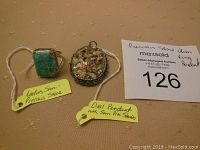 Two items displayed on tan background: a ladies semi-precious stone ring with turquoise-colored stone in gold-colored setting and an opal pendant with semi-precious stone inlays in silver-colored setting. Yellow tags identify the ring and pendant.
