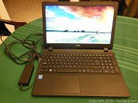 Front view of laptop powered on, screen displaying desktop background, power adapter connected at left