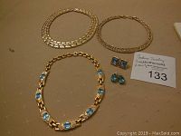 Lot of three gold-tone necklaces and two pairs of blue stone earrings arranged on a beige fabric surface with lot number tag.