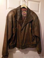 Front view of men's brown leather bomber jacket with zipper and pockets, showing jacket hanging on white door.