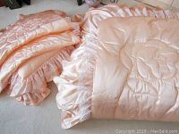 Folded double size peach satin-like bedspread with quilted floral design and lace ruffles shown folded and arranged on carpeted floor