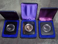 Three silver dollar coins in individual black holders inside blue velvet cases with satin interiors, front side facing.