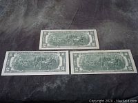 Three $2 bills laid out with their backs showing the Declaration of Independence scene.