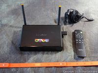 Top view of MyGica Ultra HD 4K Android TV Box with dual antennas, remote control and power adapter next to it. Orange ruler placed below for scale.