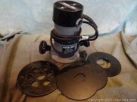 Porter Cable router with four extra base plates shown from above on gray cloth background.