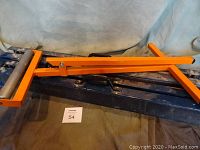 Photo showing orange adjustable height roller stand with black rollers and handle on top of black carrying case, with four folded metal folding sawhorses beneath it.