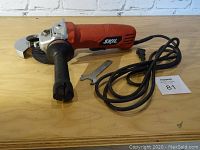 Red Skil angle grinder with attached black power cord, black side handle, metal blade changing wrench, and cut off blade with spark shield.