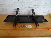 Front view of black metal TV wall mount bracket with adjustable arms and mounting bolts on wooden surface against white brick wall.