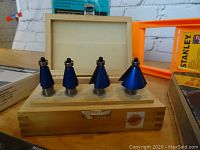 Four router chamfer bits with blue coating and steel shafts displayed in open wooden box on table