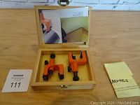 Wooden box open showing two orange magnetic jigs, instruction sheet inside box lid and additional paper on table
