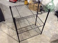 Photo of a small black metal wire shelving unit with three shelves on a concrete floor in a storage or garage setting.