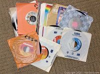 Photo showing a pile of 25 vinyl 45 RPM records in various branded paper sleeves including Columbia, CBS, WEA, and EMI.
