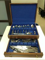 Open wooden cutlery case showing two tiers of neatly arranged cutlery including spoons, forks, knives, and serving utensils on blue felt lining.