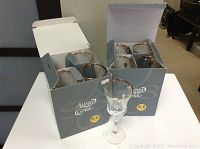 Two open boxes on a white table, each containing four clear crystal wine glasses with gold rims, one glass placed outside the box in front.