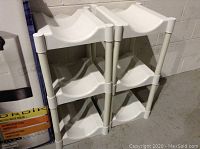 Two white plastic racks with three curved bottle slots per level, stacked side by side on a concrete floor against a wall.