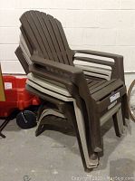 Stacked five plastic Muskoka chairs leaning against a cinder block wall in storage area. Mixed colors: two brown and three beige/tan.