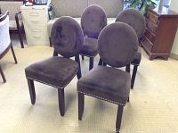 Four brown suede dining chairs arranged in a room with light-colored carpet and wooden furniture. Chairs have rounded backs and studded trim along seat edges.