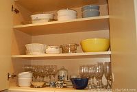 Photo of kitchen cabinet shelves holding vintage Pyrex bowls, white Corningware covered casseroles, handmade ceramic coffee set with blue floral design, clear glass wine flutes, and glasses.