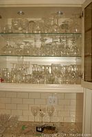 Three shelves filled with assorted clear glassware including wine, brandy, cocktail, scotch, shot, and crystal glasses arranged in a cabinet.