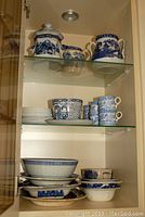 Shelf displaying various blue and white porcelain pieces including covered sugar bowl, coffee cups, saucers, and stacked plates and bowls.