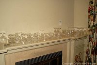 Wide view of assorted clear crystal stemware arranged on a shelf over a mantel, showing multiple glasses in different sizes and shapes, crystal decanter, and candlesticks.