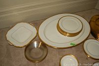 White bone china dishes with gold rims arranged on floor, includes plates, cups, and a large platter with visible crack.