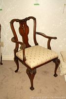 Armchair angled view showing carved wooden frame, curved armrests, patterned upholstered seat, and cabriole legs with carved feet.