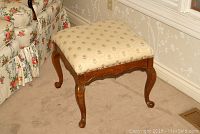 Wood stool with upholstered seat in beige floral fabric, brass nailhead trim along edge, cabriole legs, visible scratches on wood