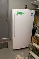 Full front view of white upright freezer with a single door, green tape on door, standing against wall.