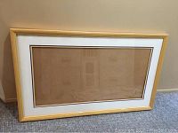 Full view of large wooden rectangular frame with glass, showing white mat with purple-yellow accent lines inside frame, placed against wall on carpeted floor.