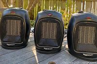 Three Amazon Basics black space heaters arranged outdoors on a wooden table, each with a visible ceramic heating element behind a front mesh grill and the brand logo printed at the base.