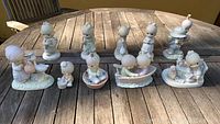 Overview image showing 10 porcelain Precious Moments figurines arranged in two rows on a wooden table outdoors. Figurines feature children in pastel colors with animals and various sweet poses.