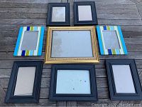 Seven easel-back picture frames arranged on wood surface showing variety of sizes and styles including black frames, colorful striped frames, and an ornate gold frame.