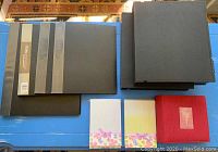 Photo showing 4 black portfolio binders stacked in two piles, 3 photo albums with floral decoration and red textured album, and 2 black protective binders.
