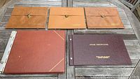 Five vintage folios shown on wooden table, including three brown leather folios with clips and two larger portfolio style folios (ledger and stock certificate portfolio).