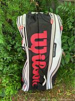 Photo showing black Wilson large tennis racket bag with prominent red Wilson lettering and white accent lines on fabric outside a green shrub background.