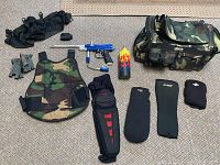 Top-down view of the paintball gear including marker, vest, gloves, knee pads, barrel cover, and camo bag.