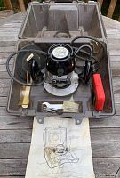 Open brown hard case showing black Craftsman router with attached power cord, wrench, small parts in plastic bags, red plastic case, and instruction sheet beneath.