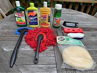 All car waxing and cleaning liquids along with brushes and polishing bonnet shown on a wooden outdoor table