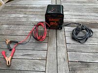 Full view of the Schauer battery charger on a wooden table showing power cord, red/black battery clips, and ventilation slots on top