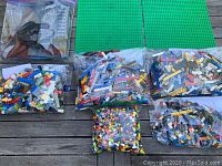 Six resealable bags filled with assorted LEGO pieces in various colors and shapes, arranged on a table with two green LEGO baseplates above them.