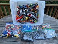 Large clear tote filled with assorted plastic building pieces with eight plastic bags containing smaller categorized pieces laid out in front on a wooden table outdoors.