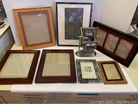 Nine assorted picture frames displayed on a white surface, varying in size, style, and material. Includes wood and metal frames; one with a floral print insert; others have glass and neutral backings or empty.