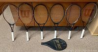 Overview of six tennis rackets arrayed on floor, showing Wilson and Prince brand rackets with new white overgrips.