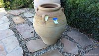 Front and side views of a large terra cotta outdoor urn with three handles placed on stone and pebble ground.