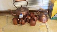 Photo showing two solid copper teapots and six matching copper cups arranged on stone floor.