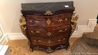 Front view of bombe chest featuring three drawers, walnut burl veneer, brass ormolu mounts, and black marble top