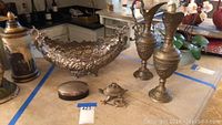 Wide view showing the silver plated decorative bowl, brass pitchers, brass vase with lid, small metal box, and brass frog figurine arranged on a table with kitchen in background.