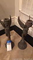 Two metal angel shaped candle holders with patina finish, seen together on floor near wall corner