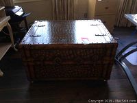 Front view of a large rectangular decorative trunk with leather covering and metal rivet accents, sitting on hardwood floor.